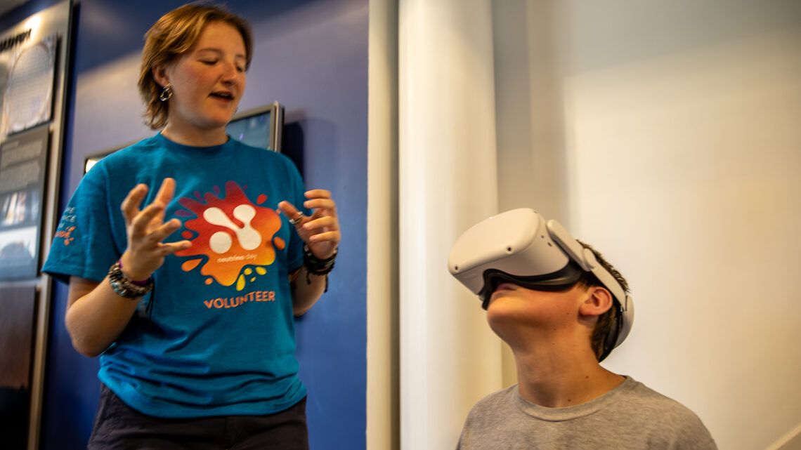 A child looks though VR goggles while a volunteer explains what they are seeing. 
