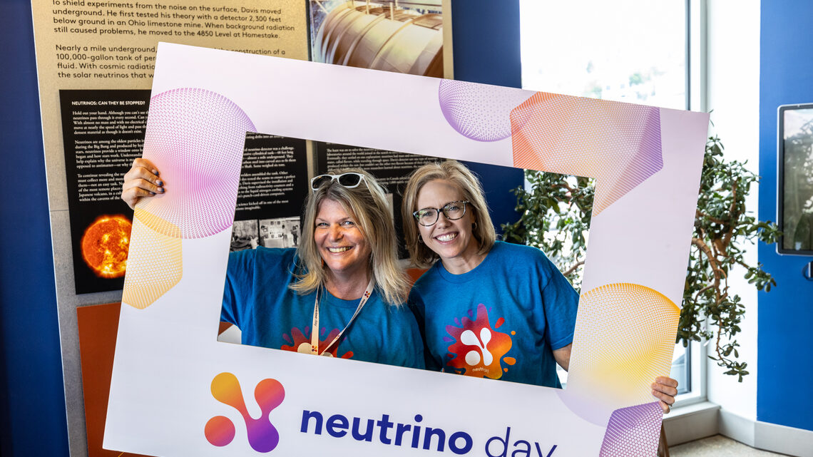 Two people smiling while holding the Neutrino Day selfie frame. 