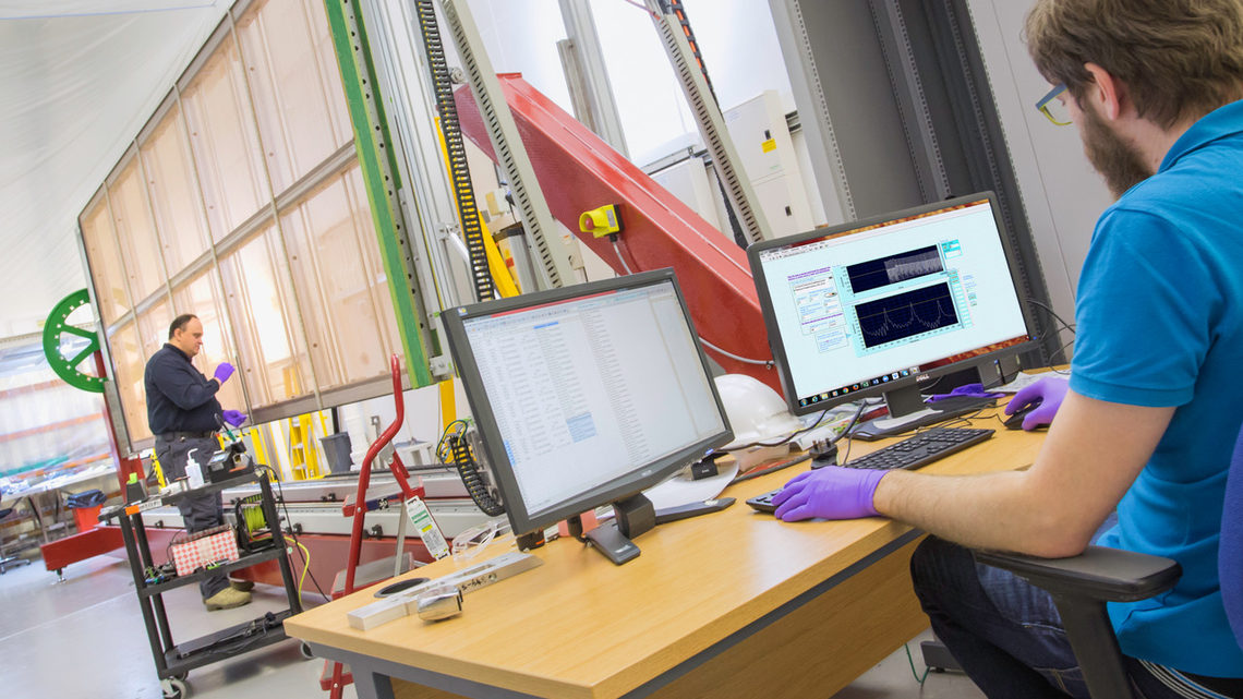 An individual in purple gloves examines the wires of the APA. A colleague, also in purple gloves, watches a computer monitor.