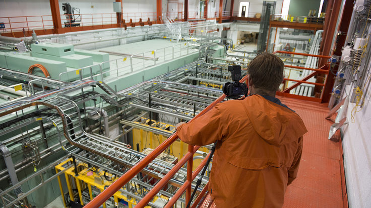 Person wearing an orange jacket takes a picture of a room full of equipment from a catwalk.