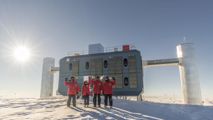 Four researchers in red coats stand on white snow at the South Pole in front of a building.