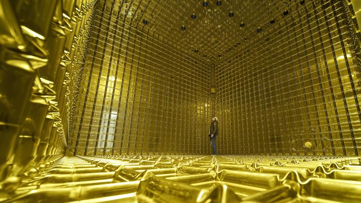 A person stands inside the ProtoDUNE detector, dwarfed by the empty space. The walls, floor, and ceiling are golden.